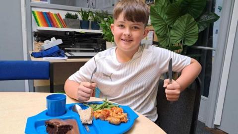 A pupil sits with a knife and fork looking like he is about to tuck into a meal and drink on a blue tray