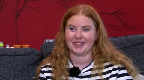 A young girl with ginger hair and a black and white horizontally-striped top smiles while sitting on a couch