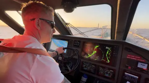 A coxswain wearing sunglasses and a white, short-sleeved shirt sits in the cabin of a fast boat with his hand on a steering wheel. Computer screens on a dashboard show navigation details. Through the front window a wide river can be seen, with the tall Grimsby Dock tower in the distance. The sky is blue and orange as the sun sets.