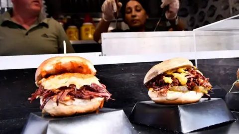 Some sort of pulled pork burger thing in the foreground and people serving it in the background