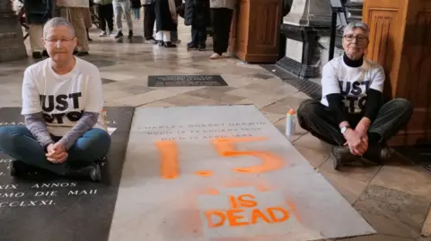 Two women wearing 'Just Stop Oil' T Shirts sitting on the ground next to Charles Darwin's stone  grave, which has '1.5 is dead' written in paint on it.