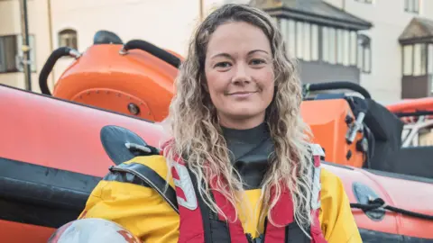 Tamara Brookes is standing in front of a RNLI rigid inflatable boat and is smiling at the camera. She has long, curly blonde hair and is wearing her RNLI uniform and buoyancy aid.