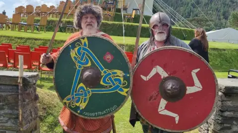 Two men with white beards dressed as Vikings with shields in front of the Tynwald Hill. One shield is green with a blue and yellow Norse design, while the other is red with the three legs of Mann in white on it.