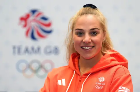 Olympic rower Becky Wilde smiles at the camera with a large Team GB logo in the background. She is wearing a branded Team GB red hoodie which also has an Adidas logo on it