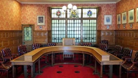 The Legislative Council chamber. A horseshoe-shaped wooden table flanked by red leather clad chairs with a larger pale upholstered chair at its head. The room has pale brown wood panelled walls and orange and yellow patterned wallpaper and includes framed old portraits on the right hand wall. There are window panels at the far end of the room with three crests in stained glass at the top.