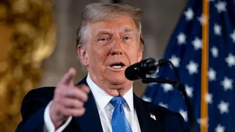 Trump at microphone with blue tie and dark suit before an American flag 