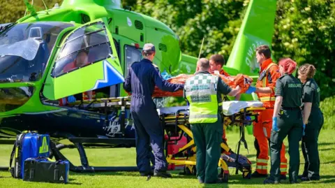 Six crew members dressed in orange hi-vis suits, blue overalls, or dark green ambulance uniforms. They are loading a mannequin on a stretcher into a green air ambulance helicopter during a training exercise in the middle of a field.