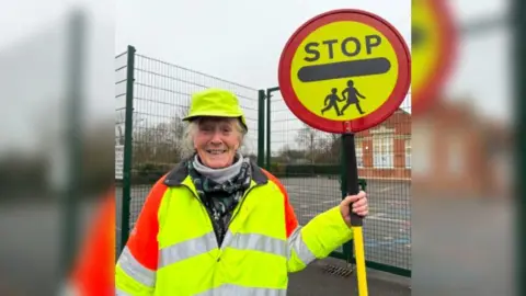 Burnham-on-Sea.com Heather standing outside on a dull cloudy day in her high vis jacket and holding her lollipop stick, in front of a school playground