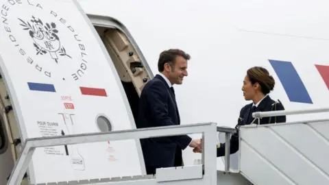 French President Emmanuel Macron stepping off plane in Mayotte, greeted by a woman 