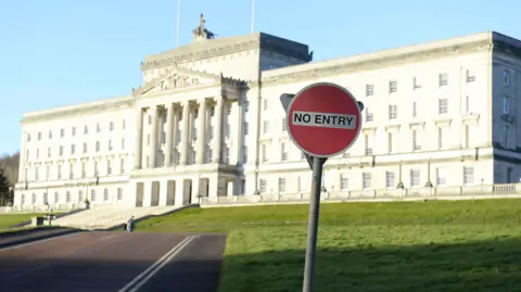 Parliament Buildings at Stormont, a large and elegant building of white brick, with columns to the front. In the foreground is a traffic sign with the words No Entry written on it.
 