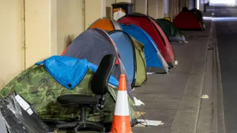 A row of pop-up tents on a street pavement with rubbish, including an old office chair and a traffic cone in the foreground