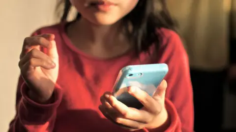 Getty Images A stock image of a young child holding a pale blue smartphone. She has long dark hair to her shoulders and wears a red top with sleeves.