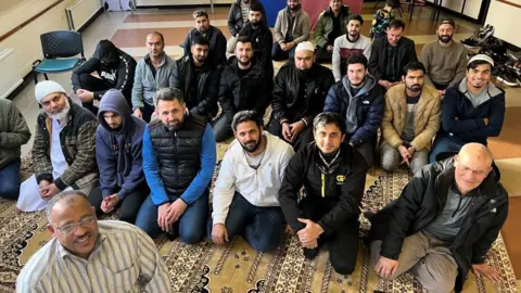 "Mobile imam" Mohamed Osman and worshippers at Selby Friday prayers. Mohamed is at the front of the picture, smiling, wearing spectacles and a stripy shirt. The other worshippers are kneeling behind him on the prayer mat.