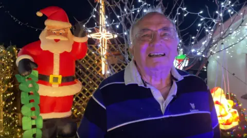 BBC Mr Langman is stood in his garden which has been decorated with Christmas lights. Mr Langman is wearing a black and blue striped Guinness shirt. He has short hair and wears glasses. He is stood in front a small tree which has been decorated with fairy lights. A large inflatable Father Christmas decoration has been placed behind Mr Langman.