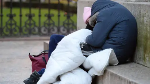 Person huddles on a step wearing warm coat and pink hat trying to keep warm, with a duvet round them. A red canvas bag lies on the floor.