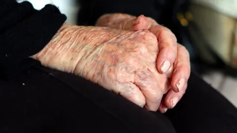 The hands of an elderly woman at home. She is wearing black and has chipped pink nail varnish on her fingers. 