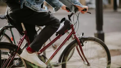 A stock image of a man cycling on the pavement. You can see he is on a burgundy colour bike frame, with a street and black lamppost in the background. He is wearing black skinny jeans with red socks and white trainers, with a light blue denim jacket.