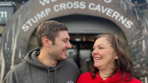 BBC/ Julia Lewis Oliver, who wears a grey hoodie, and his mum Lisa, who wears a red hoodie, look at each other and smile outside a building with a sign reading "Stump Cross Caverns".