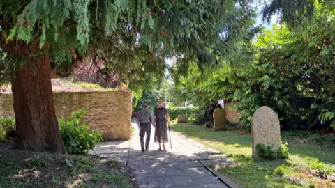 sunseeker WEDNESDAY - A man and woman hold hands walk through a graveyard on a sunny day. They are both wearing straw hats. The woman is using a walking stick and is wearing a dress, the man is wearing a shirt and trousers. They are walking under a pine tree into the sunshine. On their left is a brick wall, on the right are several gravestones and a tall green hedge.