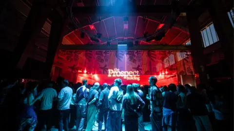 The bar area at the Prospect building. The room is dark with red and blue lights. There is a crowd of people standing around.