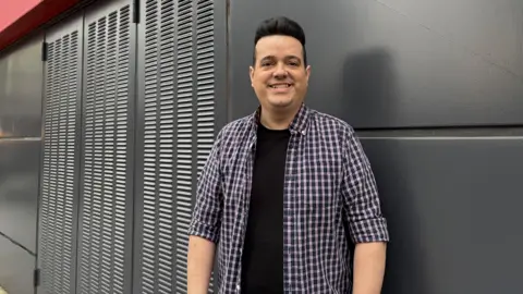 Vinny Ferrer is standing in front of the dark grey exterior of the new shop. He has black hair styled in a quiff and is wearing a black t-shirt with a blue, purple and white check shirt over the top.