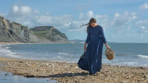 Andrew Blackmore Katy Hamilton in blue Victoria-era dress on Lyme Regis beach with cliffs and sea in the background