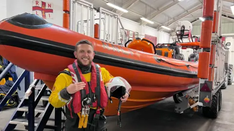 Connor Wray Connor Wray in his yellow lifeboat volunteer uniform and red lifebelt, holding a helmt and standing in front of an elevated orange RNLI boat in its station depot