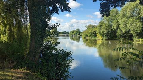 The River Thames at Wallingford looking towards Benson Lock with trees either side and a blue sky with clouds reflected in the still water
