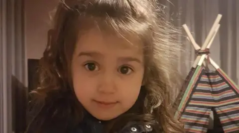 A handout image shows Sara Sharif, a 10-year-old girl whose father and stepmother have been convicted of her murder. At the time the image was taken, she was four. She had long brown hair and is seen looking into the camera. 