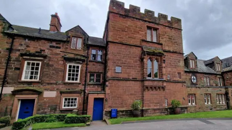 The Prior's Tower by Carlisle Cathedral. The three-storey structure is red sandstone and has windows on the first and second floors. It is flanked by the other buildings also made of sandstone.