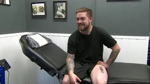 Dan Thomas sitting on a tattoo couch holding the leg of his shorts up to show the tattoo, which is the Euros cup with the words: "England Euro 2024 Winners"