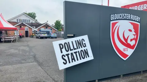 BBC The Lion's Den with a red sign near 'polling station' sign with building and car park behind.
