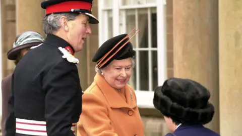 Pacemaker An archive shot of Elizabeth II in 2000, she is wearing an orange coat and a black and orange hat. To her right is a tall man in a dark, formal military uniform, to her left is a woman, seen from behind, wearing a dark blue jacket and fur hat.