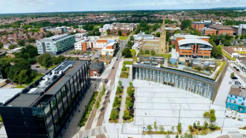 University of Central Lancashire drone view of a modern multi-storey black building at the left of a pedestrian plaza with rows of shrubs and other multi-storey buildings and an old church in the background