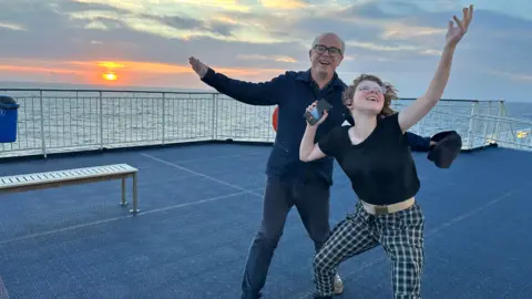 Julian Francis Julian Francis and April, smiling and throwing their arms in their air on board a ferry at sunset. Mr Francis wears black-rimmed glasses, a navy shirt and jeans. April wears glasses, a black t-shirt and chequered trousers, holding a phone in one hand.