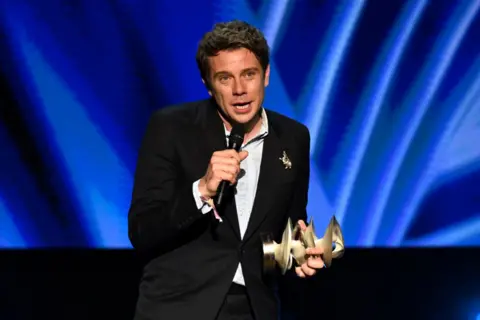 Jonathan Anderson in black suit and white shirt, holding microphone as he accepts gold award against a blue backdrop