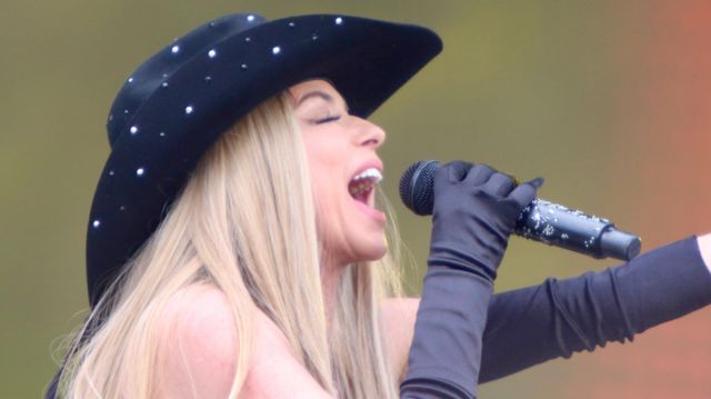 Shania Twain wears black gloves and a black cowboy hat while singing into a microphone