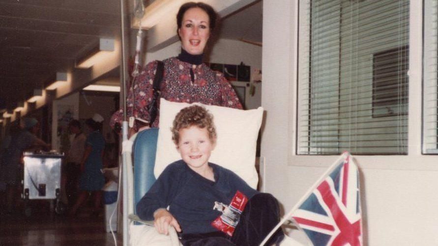 Antonya Cooper stands behind her young son Hamish who sits in a hospital chair.