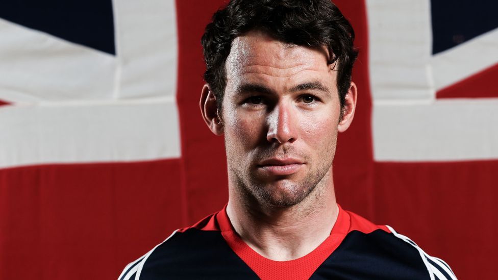 Mark Cavendish, with a serious expression and wearing a blue top with red and white piping, stands in front of a Union Jack