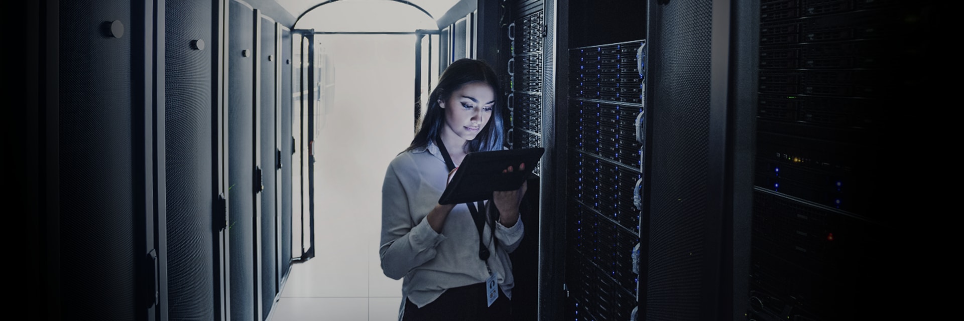 An IT professional reviews data on a tablet, surrounded by dark server racks illuminated by small LED lights in a high-tech data center