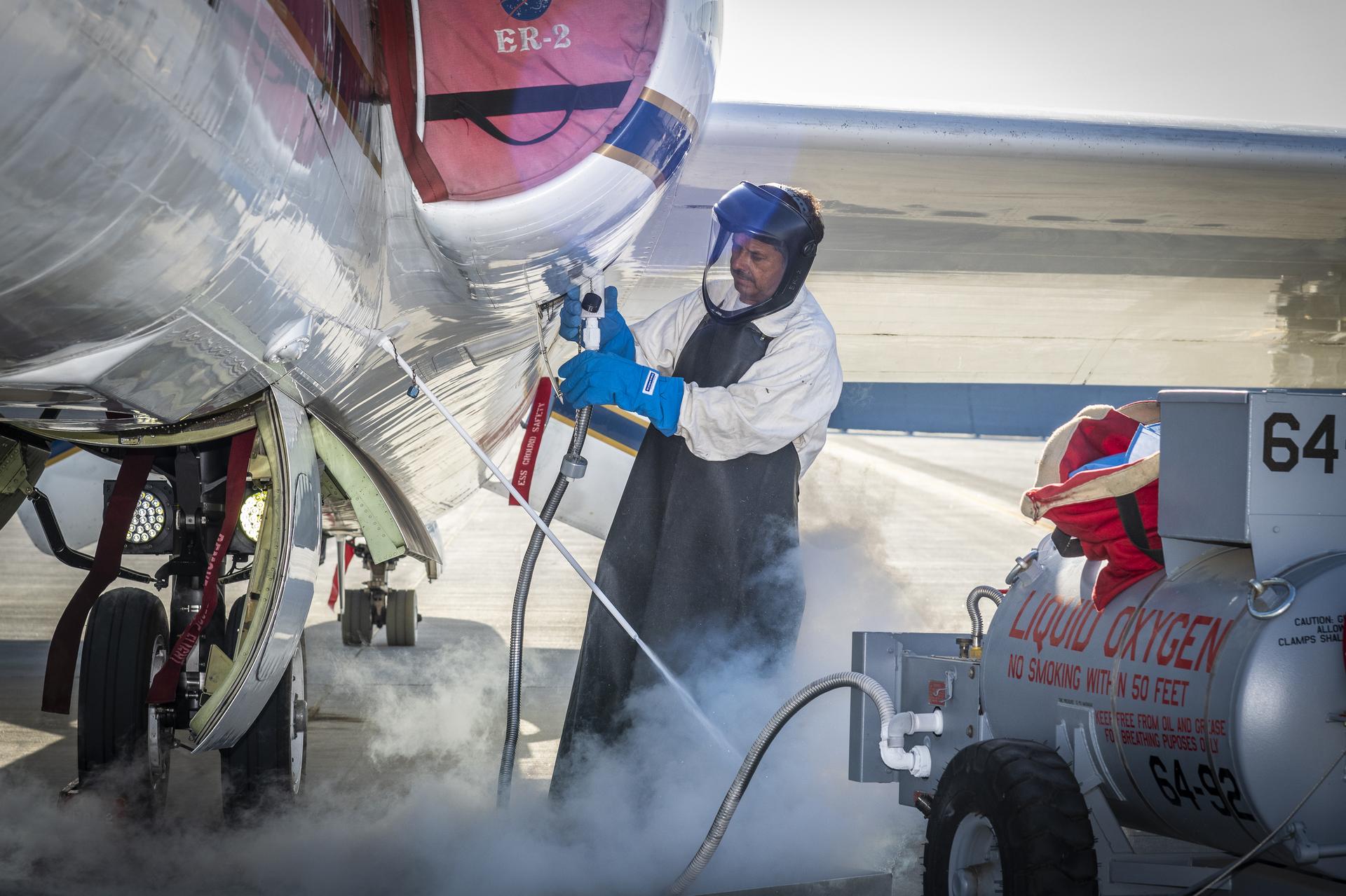 A man clad in a white protective suit, black apron, and blue gloves administers liquid oxygen from a tank to a parked aircraft via hose. Gas clouds hover near his feet and the man is wearing a helmet with a clear visor over his face. 