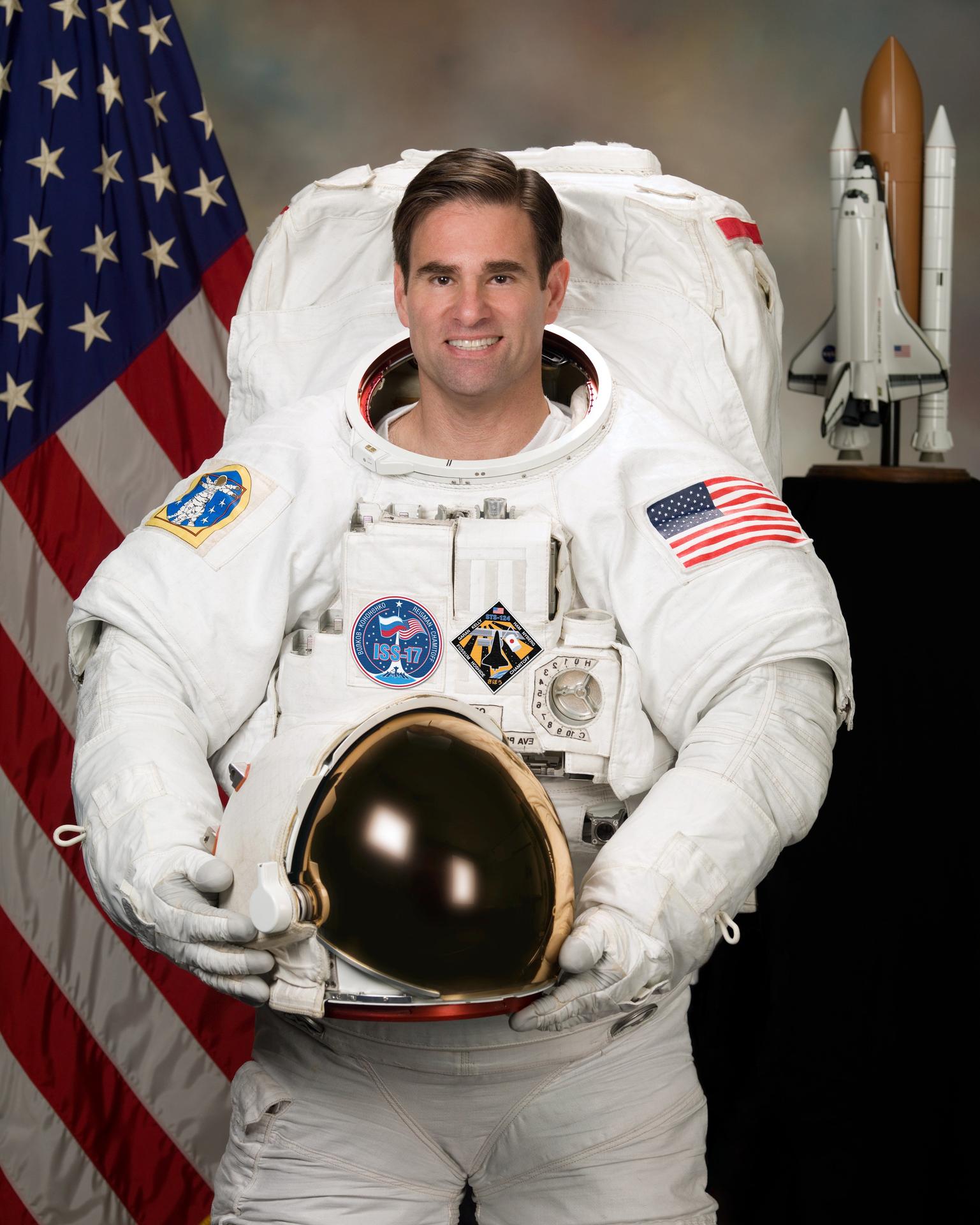 A man wearing a spacesuit smiles and stands in front of the American flag. He is holding his helmet in front of himself.