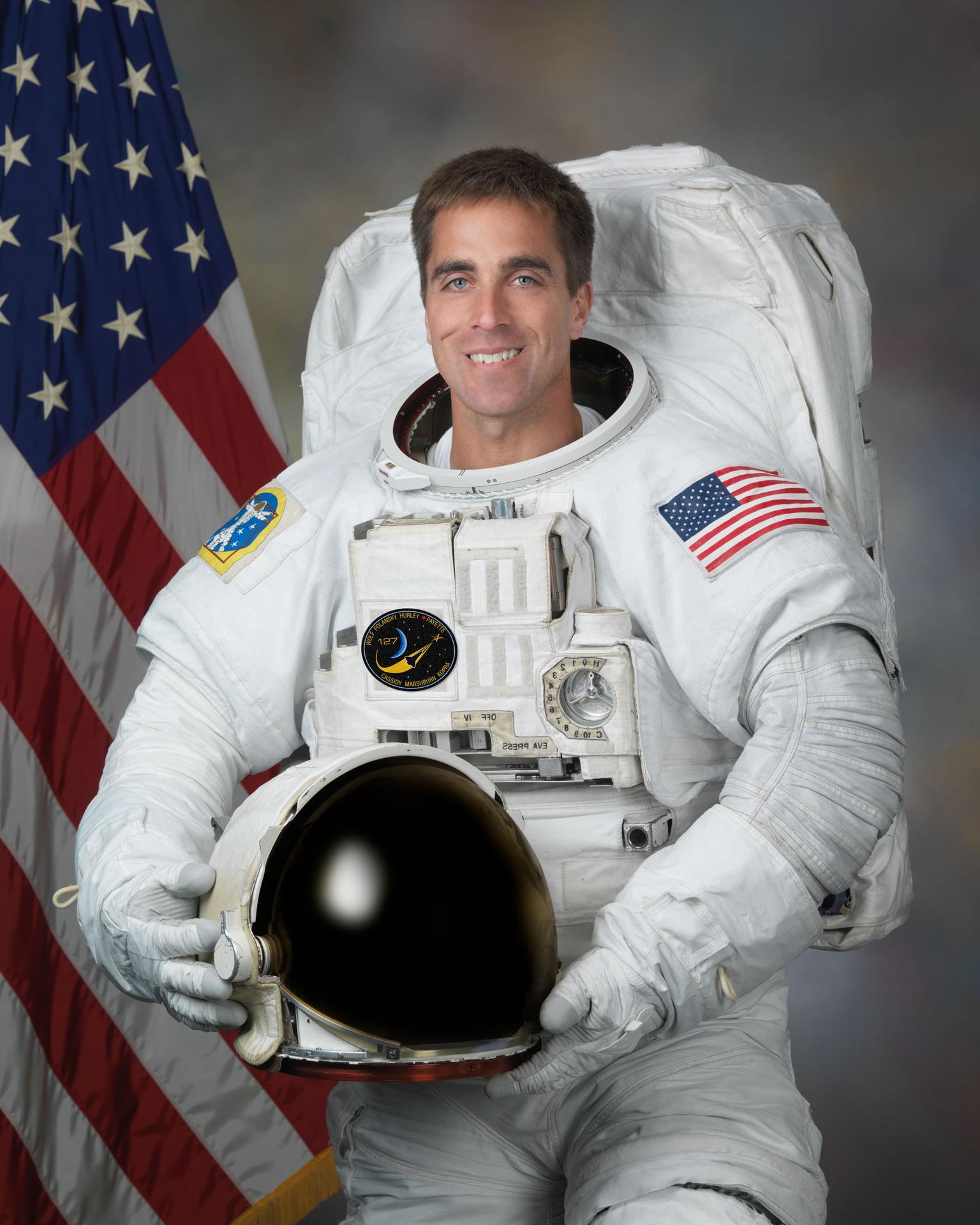 A man wearing a spacesuit smiles and stands in front of the American flag. He is holding his helmet in front of himself.