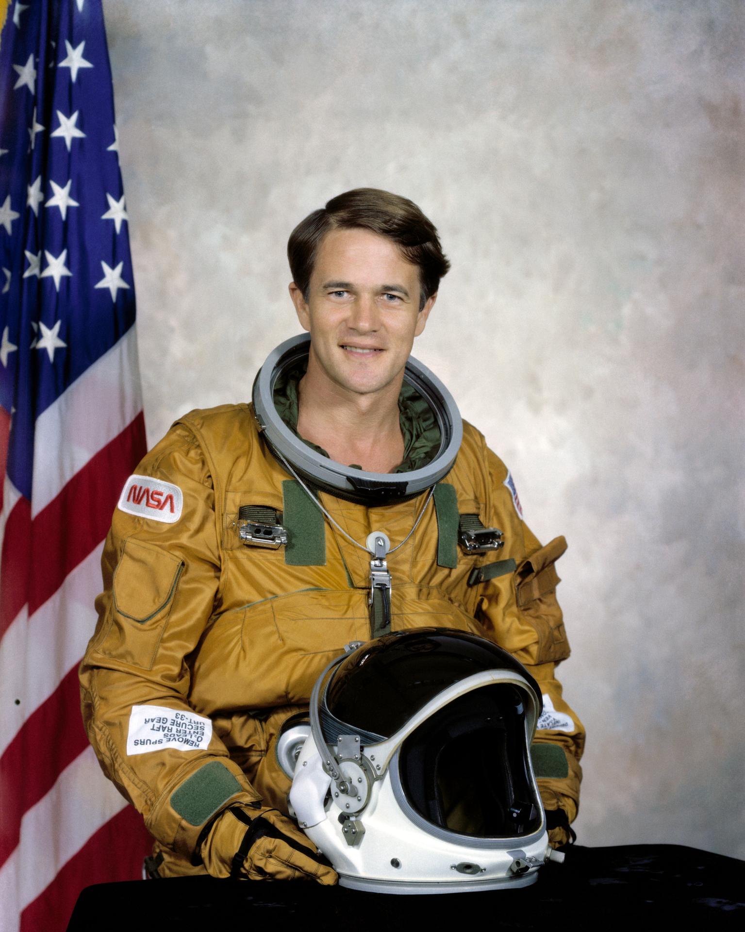 Man sitting in an astronaut suite, with his helmet off and holding it in his lap.