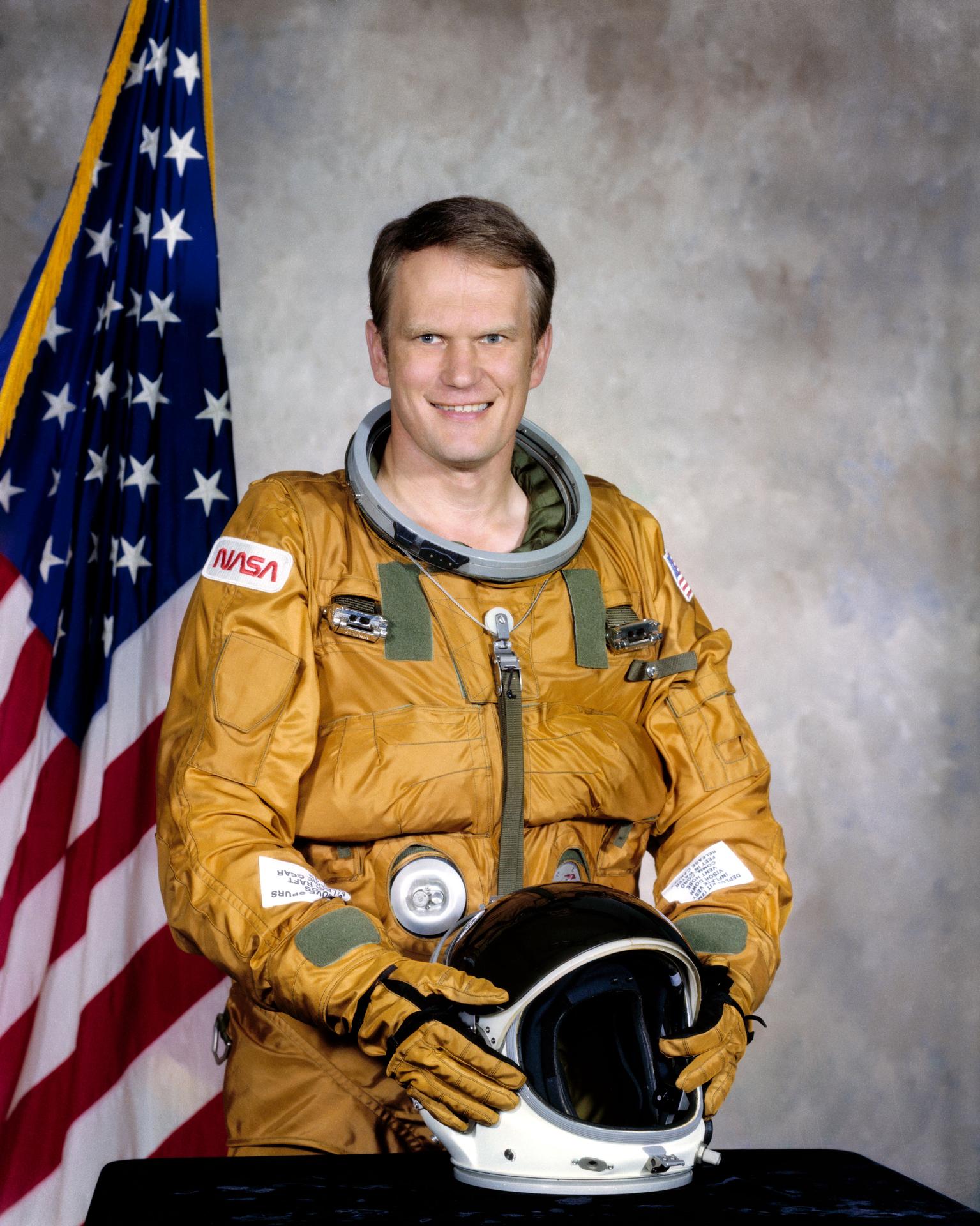 A man wearing a spacesuit smiles and stands in front of the American flag. He is holding his helmet in front of himself.