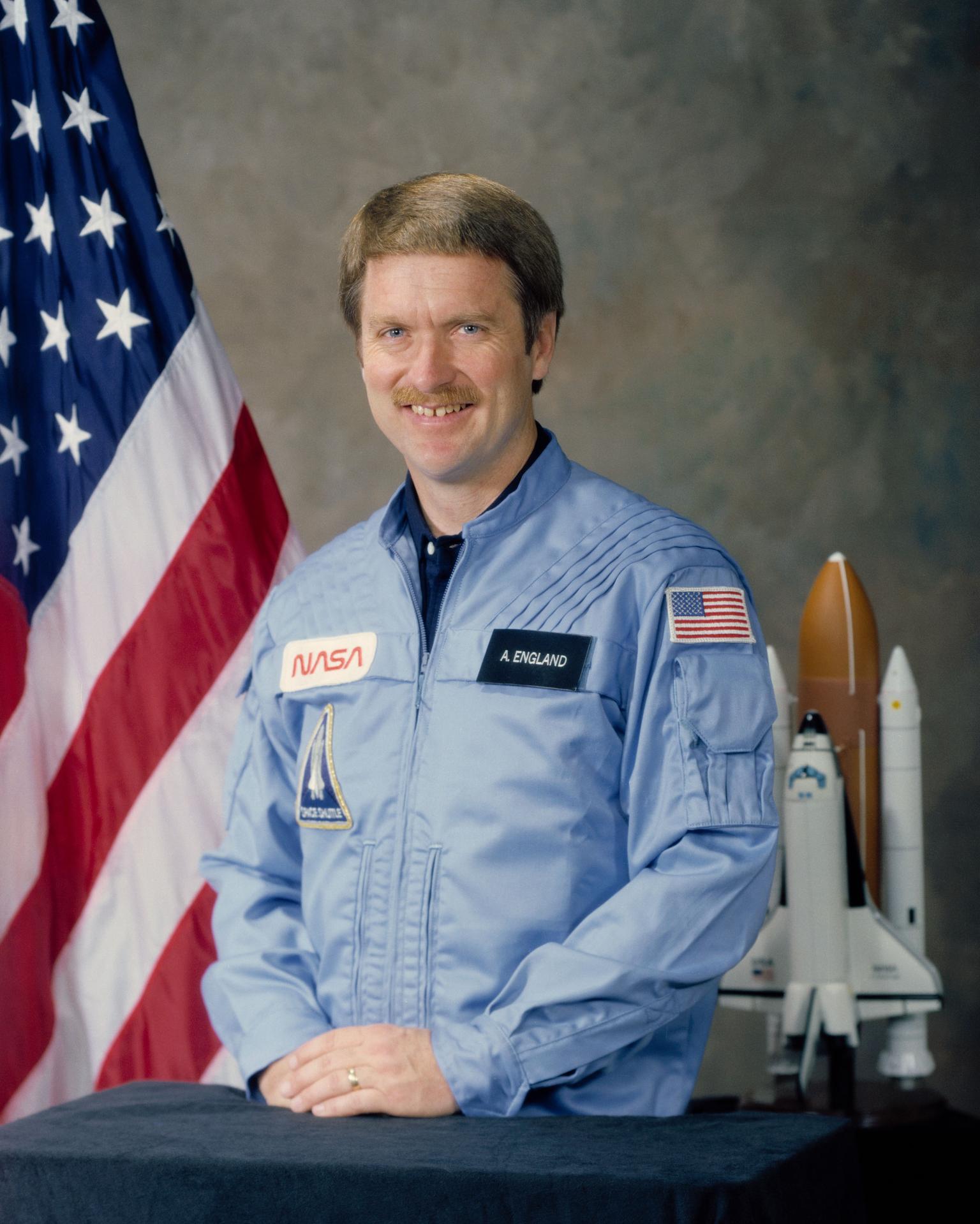 A man wearing a space flight jumpsuit sits and smiles. There is an American flag behind him and a small module of a rocket ship.