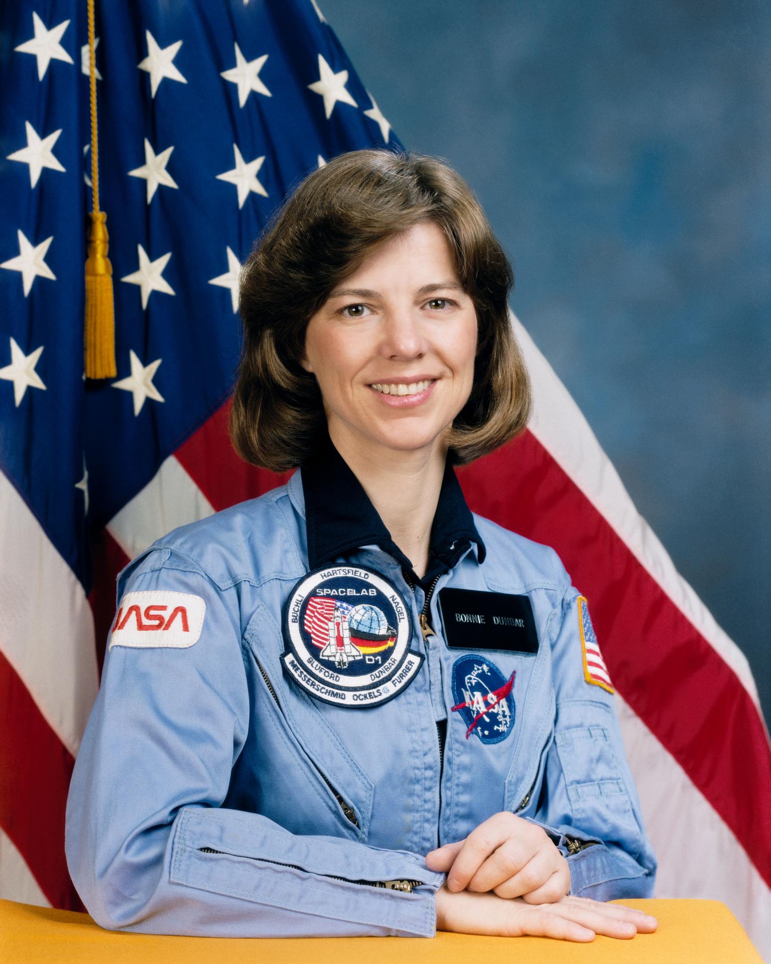 A woman with shoulder length hair smiles wearing a NASA flight suit, holding a her hands on a table before herself.