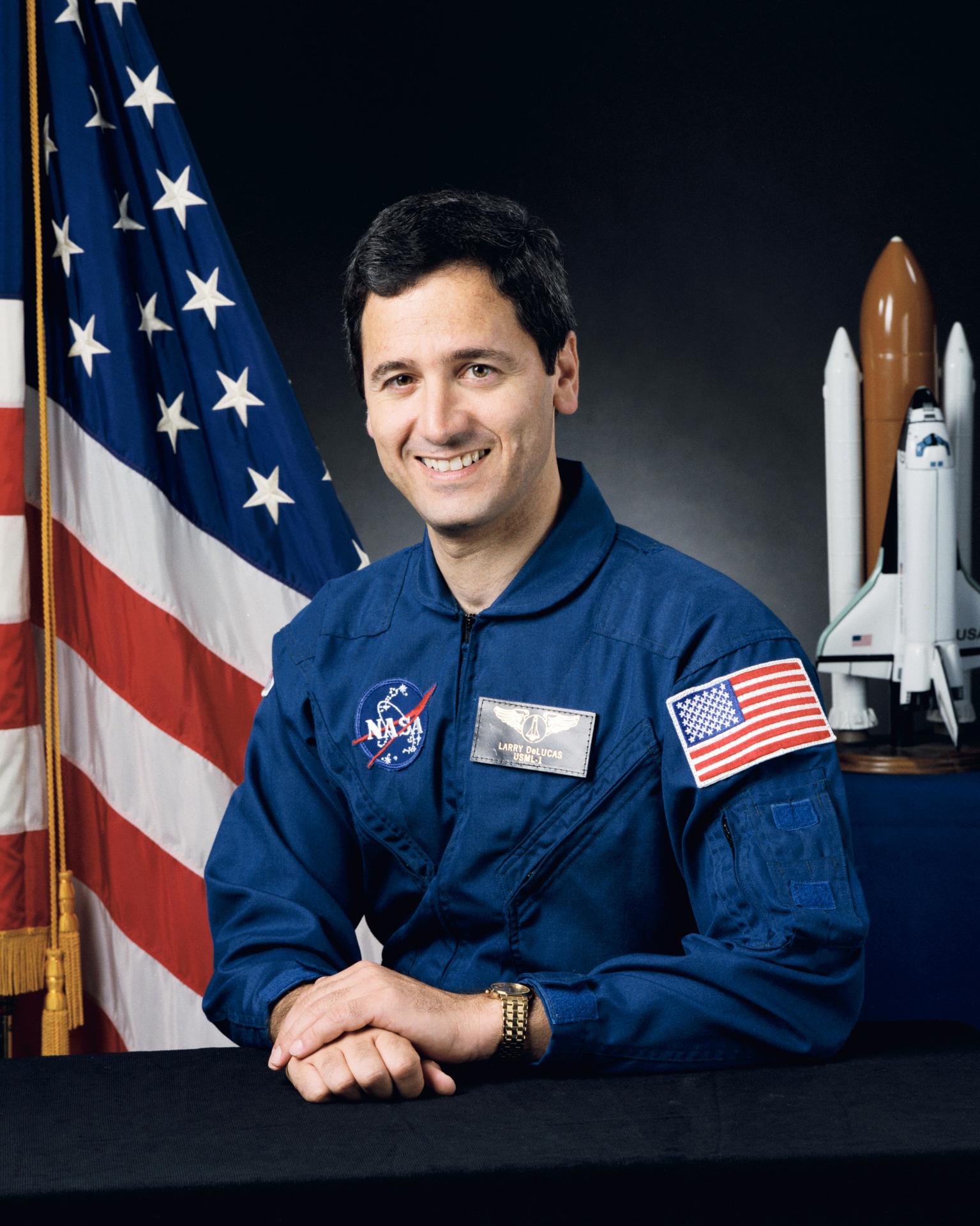 A man smiles, wearing an astronaut flight suit. He is sitting in front of the American flag and beside a model rocket.