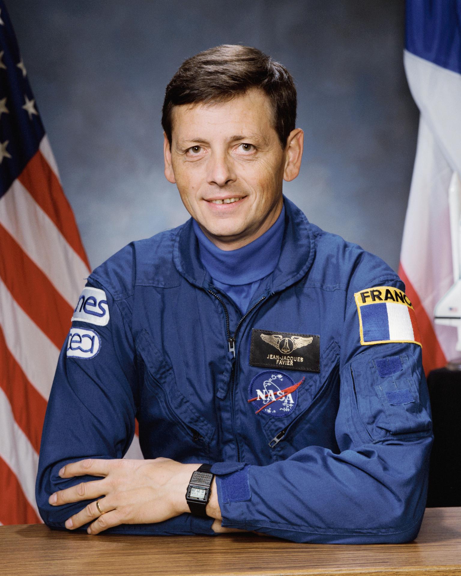 A man wearing a space flight jumpsuit sits and smiles. There is an American flag behind him.