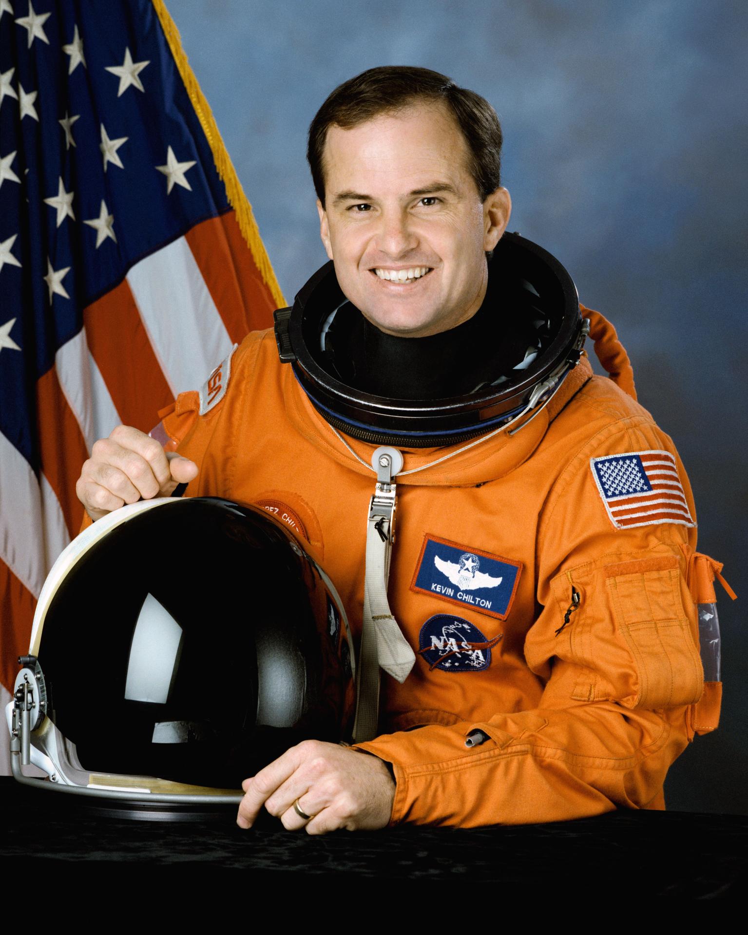 A man wearing a spacesuit smiles and stands in front of the American flag. He is holding his helmet in front of himself.